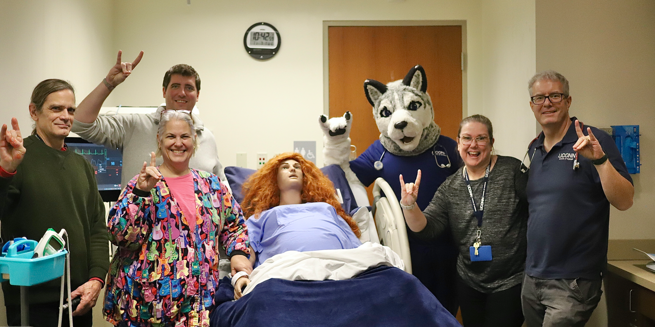 Sim lab faculty making husky sign and standing around a manikin and with mascot Jonathan the Husky