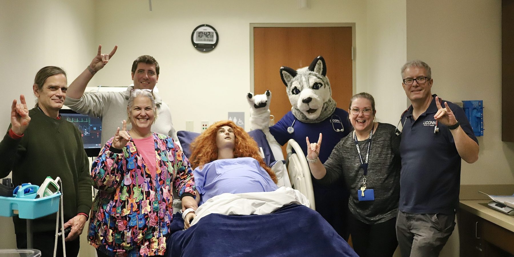 Simulation Lab faculty making Husky sign with their hands standing around a manikin and with the mascot Jonathan the Husky