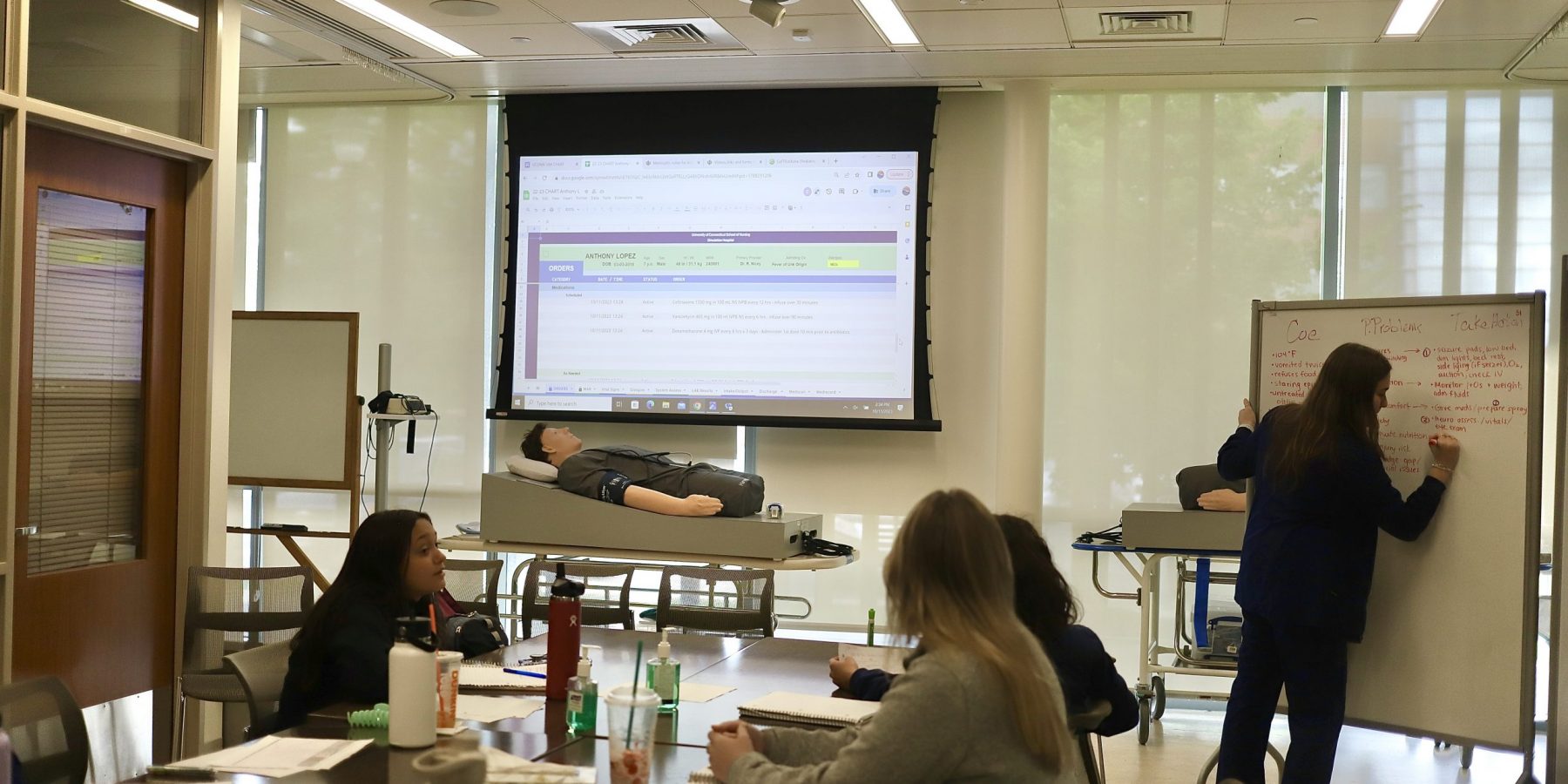 Student writing on whiteboard in front of sim lab and three students watching